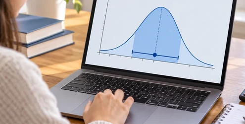 A student studies a bell curve with a highlighted confidence interval on a laptop beside a calculator and notebook.