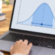 A student studies a bell curve with a highlighted confidence interval on a laptop beside a calculator and notebook.