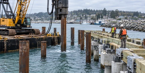 A barge-mounted pile driver sets foundation piles for a new harbor pier while marine construction workers in flotation gear monitor the work.