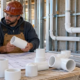 An engineer and plumber compare PVC pipes and fittings with a service drawing beside separate drainage pipe and conduit runs.