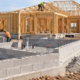 Builders progress from a concrete masonry foundation to timber framing while materials are checked on site.