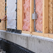 Engineer checks a framed wall assembly with insulation, weather barrier and services above a concrete foundation.