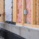 Engineer checks a framed wall assembly with insulation, weather barrier and services above a concrete foundation.