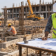 A construction manager and civil engineer review site plans as crews tie reinforcing steel and an inspector checks footing layout at an active building project.