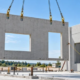 Crane lowers a precast concrete wall panel while workers guide it into position from a safe distance.