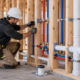A plumber installs supported supply and drain lines through open house framing before the walls are closed.