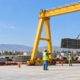 Workers guide a bundled steel-bar load lifted by an overhead gantry hoist inside a marked exclusion zone.