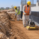 Workers compact a soil backfill lift beside a waterproofed foundation wall with a drainage layer.