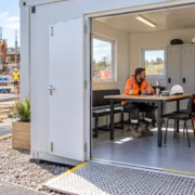 Construction workers use a clean site welfare cabin with handwashing, drinking water and rest seating beside a portable toilet and active build site.