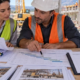 An accountant and project engineer track current construction costs against progress plans while the unfinished building remains active on site.