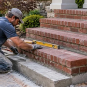 A mason levels brick stair treads on a prepared base while a supervisor checks the stair alignment and mortar joints.
