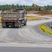 Compacted gravel access road lets a loaded dump truck enter a construction site beside a drainage ditch.