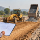 Engineer reviewing road project costs beside a grader, aggregate truck, and asphalt paver