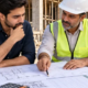 Homeowner and contractor compare detailed house plans, construction cost estimates, and material samples at a residential jobsite