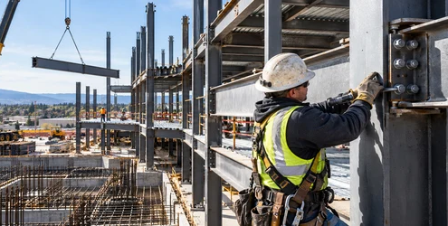 Steel erector bolts a structural beam into a building frame as a crane lifts another steel member.