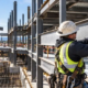 Steel erector bolts a structural beam into a building frame as a crane lifts another steel member.
