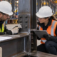 Steel fabricator and inspector check a structural beam connection and weld against shop drawings in a fabrication workshop.