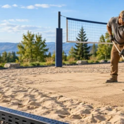 Workers rake and level the sand court beside a volleyball net and drainage edging during court construction.