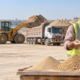 Contractor and supplier compare sand and aggregate samples beside bulk stock and a delivery truck to assess construction supply logistics.