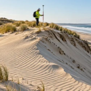 Field engineer surveys a wind-shaped coastal dune with native grass and sand fences protecting the shoreline.