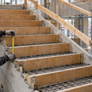 A carpenter measures the vertical riser formwork on a reinforced concrete stair while an engineer checks the landing levels.