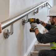 A carpenter secures a wall-mounted stair handhold bracket while an inspector checks the grip rail along the stair.