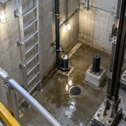 An engineer checks the fixed ladder and sump pump access inside a guarded elevator pit with the rails and drain visible.