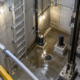 An engineer checks the fixed ladder and sump pump access inside a guarded elevator pit with the rails and drain visible.