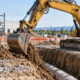 A hydraulic excavator digs a service trench while a supervisor checks the plan from outside the machine work zone.