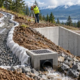An engineer checks a gravel-filled interceptor drain across a sloped construction site, carrying runoff away from a retaining wall to a catch basin.