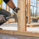 A carpenter drives an angled nail through a wall stud into the bottom plate while an engineer checks the framing detail.