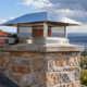 Roofer secures a stainless steel chimney cap over a masonry chimney to keep rain and debris out.