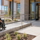 A wheelchair user follows a guarded entrance ramp toward a level landing while an accessibility consultant and engineer review the door connection.
