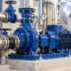 An engineer checks a commercial water pump's pipe connections and pressure gauge while a technician verifies service clearance.