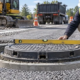 A civil engineer checks a cast-iron manhole frame with a straightedge and survey staff while a worker adjusts its bedding to match the road grade.