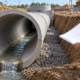 Cutaway drainage pipe in an excavation showing water at the bottom interior invert while an engineer reviews plans.