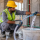 Construction plumber flushing a newly installed water line through a clear hose into a bucket before system commissioning