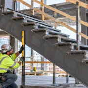 A stair installer checks a steel stringer and tread supports against the landing connection while a supervisor reviews alignment.