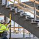 A stair installer checks a steel stringer and tread supports against the landing connection while a supervisor reviews alignment.
