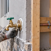 A plumber fits an exterior hose bib while an inspector checks the pipework and pressure gauge inside the unfinished wall.
