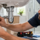 A plumber fits a metal scutcheon plate around a bathroom supply pipe at the tiled wall.