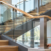 An installer fits a glass balustrade panel beneath a continuous timber banister rail while an engineer checks the newel post anchorage.