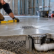 A plumber uses a level to check a sloped concrete floor leading to a fitted drain grate and below-slab drain pipe.