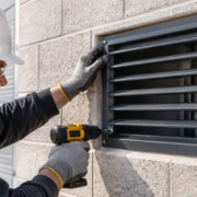 Installer secures a metal ventilation grille over a masonry wall opening on a building site.
