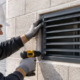 Installer secures a metal ventilation grille over a masonry wall opening on a building site.