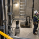 Protected service lights illuminate the guide rails, buffers, fixed ladder, and sump in a secured elevator pit.