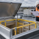 A roofer checks the curb and hinged access hatch on a guarded roof opening while an engineer reviews the installation detail.