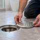 A plumber positions a removable floor cleanout cover beside the open drain access and cleaning cable.