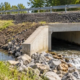 Reinforced concrete box culvert carries stream water beneath a road embankment, with riprap protecting the outlet.