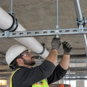 Construction worker tightening a slotted steel strut channel hanger that supports insulated pipe and conduit beneath a concrete ceiling.